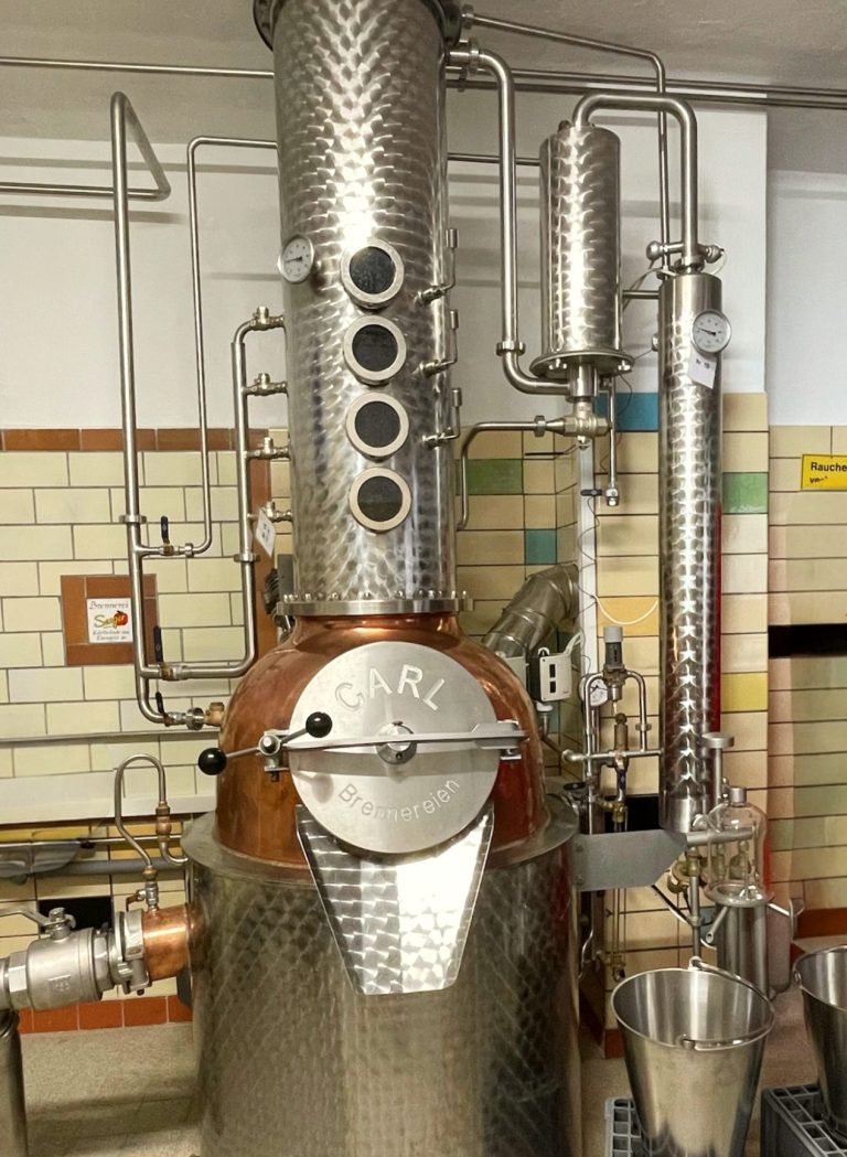 Edelstahl-Destillieranlage mit Kupferdetails und verschiedenen Rohren und Ventilen.